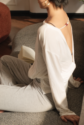 A woman sits barefoot on the floor with an open book, leaning back against the seat of a soft grey sofa in warm afternoon light.