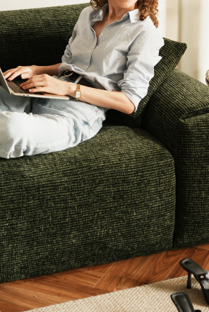 A woman works on a laptop, curled up sideways against the arm of a deep green textured sofa, her shoes kicked off on the floor beside her.